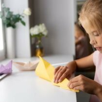 Origami, fetita cu hartie galbena la birou langa fereastra